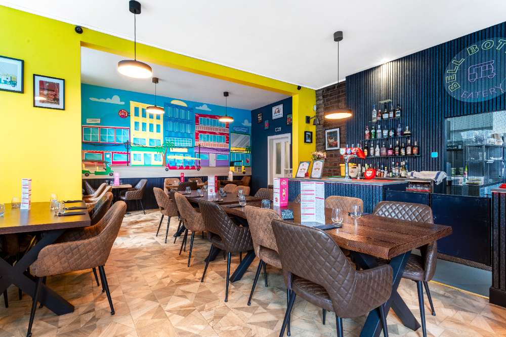 The Bell Bottom Eatery dining room — chartreuse-yellow walls, the Delhi street mural, the navy slat bar with the neon Bell Bottom sign